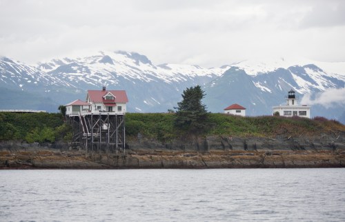 A coastal scene featuring a house on stilts, surrounded by greenery and mountains with snow-capped peaks in the background.