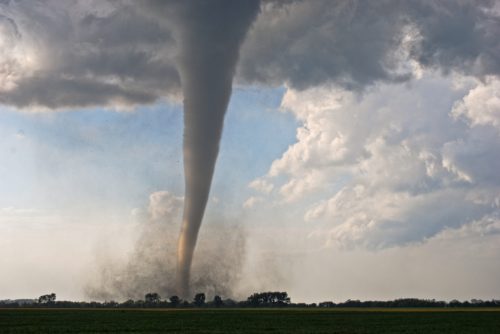 A tornado touches down in a rural landscape, surrounded by dark clouds and a clear blue sky.