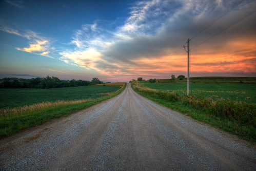 A gravel road stretches into the distance, flanked by green fields under a colorful sunset sky.
