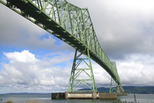 A green steel bridge spans over a calm body of water under a cloudy sky.