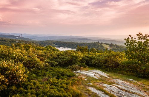 A scenic view of rolling hills and a lake under a colorful sunset sky, surrounded by lush greenery.