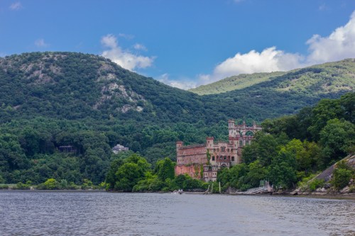 A historic castle nestled among lush green hills by a river under a partly cloudy sky.