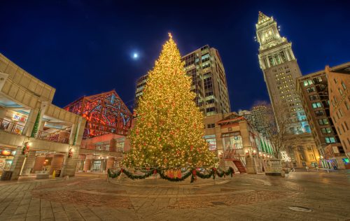 A brightly lit Christmas tree stands in a plaza at night, with a tall clock tower and festive decorations in the background.