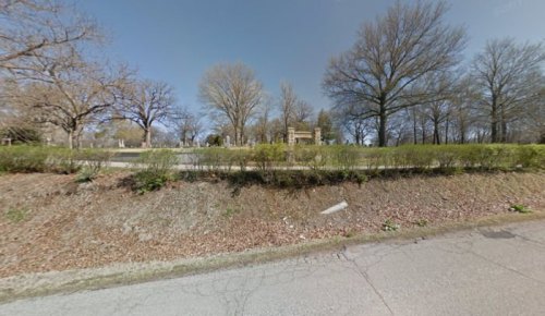 A view of a park with bare trees and a grassy area, featuring a sign in the background and a paved road in the foreground.
