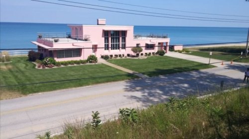A pink beachfront house with a manicured lawn, located near a road and overlooking the ocean under a clear blue sky.