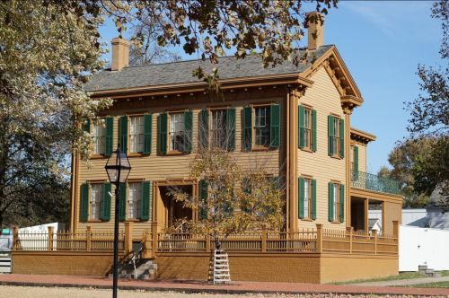 A historic two-story house with green shutters, a porch, and a landscaped yard on a sunny day.
