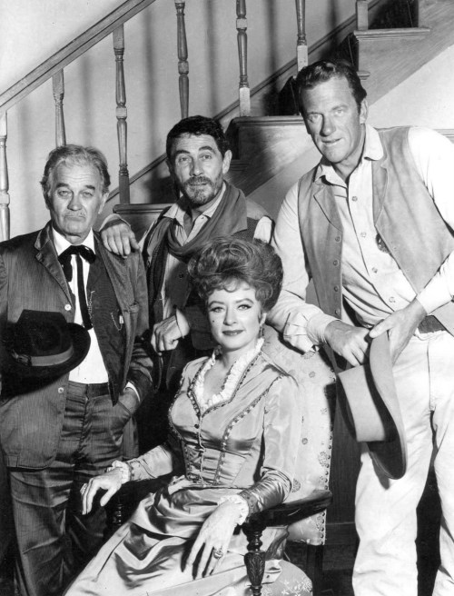 A black-and-white photo of four actors posing together, with a staircase in the background. They are dressed in vintage attire.