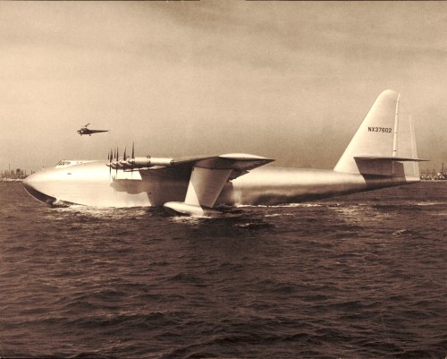 A large seaplane with multiple propellers takes off from the water, while a helicopter flies above it.