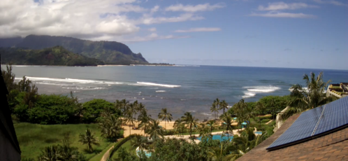 A scenic coastal view featuring waves, palm trees, and mountains under a clear blue sky.