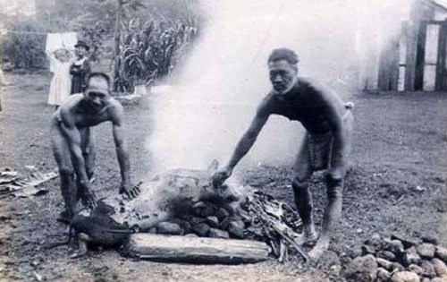 Two men preparing food over a fire, with smoke rising, in a historical outdoor setting.