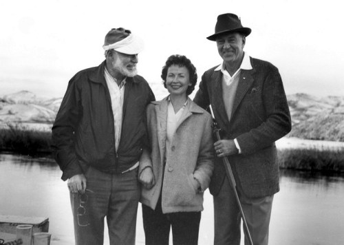 A black and white photo of three people smiling together outdoors, with a scenic landscape in the background.