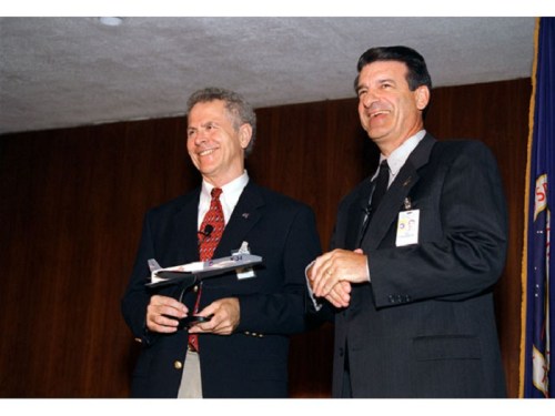 Two men in suits smile while holding a model airplane at a podium, with a flag in the background.