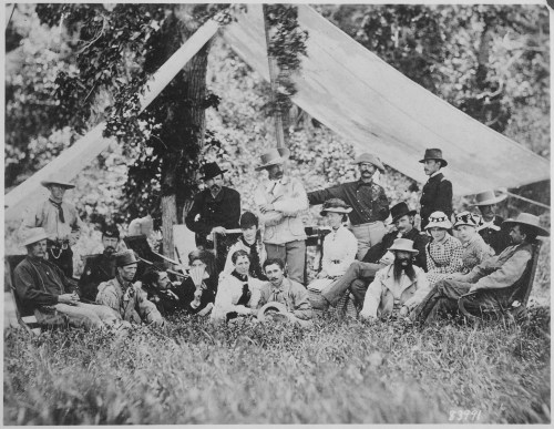 A historic black-and-white photo of a large group of people, seated and standing, under a tent in a grassy area.