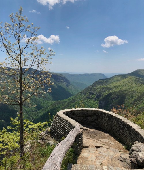 A stone overlook with a wooden railing, surrounded by lush green mountains and a clear blue sky.