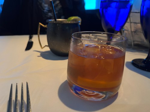 A close-up of a glass of amber-colored drink on a table, with a metal cup and blue glass in the background.