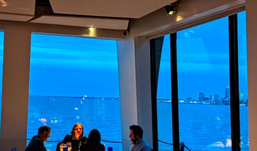 A group of people dining indoors with a view of a blue evening sky and city skyline over the water.