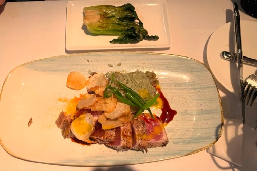 A plate with sliced meat, crispy chips, green sauce, and garnishes, alongside a small dish of sautéed greens.