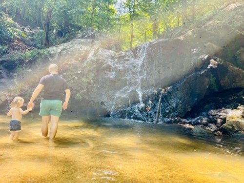 A person and a child wade through a shallow stream near a rocky waterfall, surrounded by lush greenery.