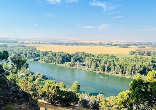 A serene river flows through lush greenery, surrounded by fields under a clear blue sky.