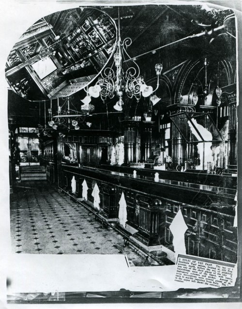 Historic black-and-white photo of a bar interior with ornate chandeliers and a wooden counter, featuring ghostly white figures.