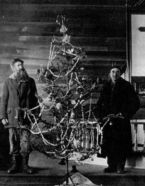 Two men stand beside a decorated Christmas tree in a rustic interior, showcasing vintage ornaments and garlands.
