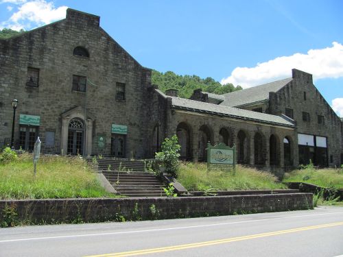 Abandoned stone building with arched windows and overgrown grass, set against a blue sky and green hills.