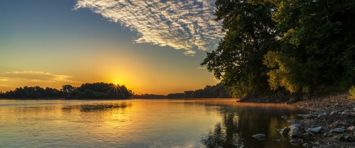 Sunrise over a calm river, with trees lining the shore and clouds reflecting in the water.