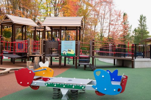 Colorful playground equipment with a seesaw, surrounded by wooden play structures and autumn foliage in the background.