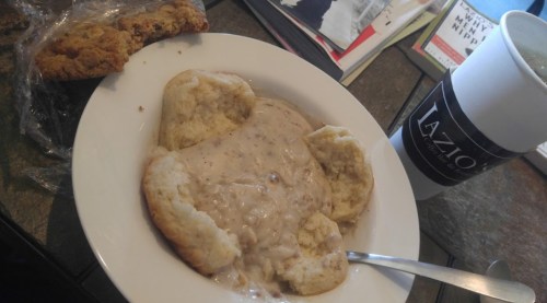 A bowl of biscuit and gravy with a spoon, alongside a cookie and a coffee cup on a table.