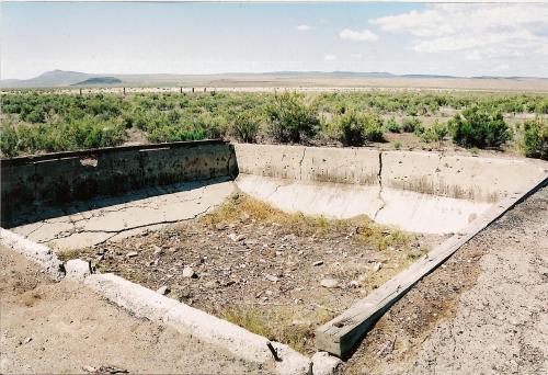 A concrete foundation sits in a barren landscape, surrounded by sparse vegetation and distant hills under a clear sky.