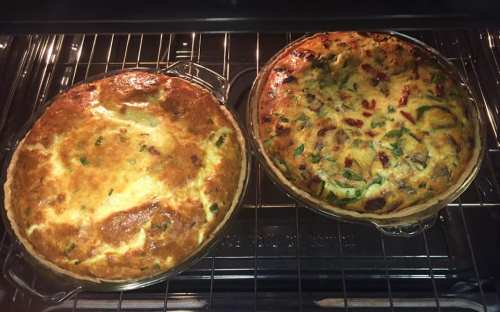 Two freshly baked quiches in glass pie dishes, one with a golden crust and the other with colorful vegetables, in an oven.
