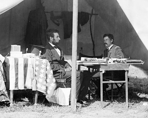 Two men seated at a table inside a tent, with an American flag draped over the table, engaged in conversation.
