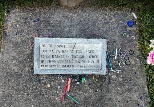 A grave marker for Louiza Catharine Fox, aged 13, murdered in 1869, with colorful trinkets and flowers around it.
