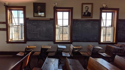 Historic classroom with wooden desks, chalkboards, and portraits on the walls, featuring large windows.