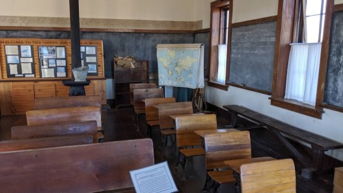 Historic classroom with wooden desks, a chalkboard, a map, and educational displays on the walls.