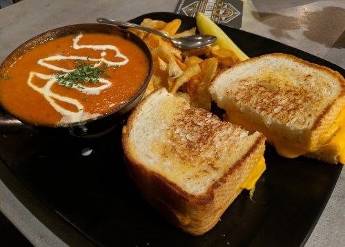 A bowl of tomato soup with a swirl of cream, served with a grilled cheese sandwich and potato chips on a black plate.