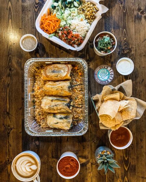 A table spread with a tray of enchiladas, rice, salad, chips, salsa, and a latte, all on a wooden surface.