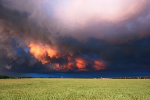 Dramatic clouds with orange and purple hues loom over a green field under a twilight sky.