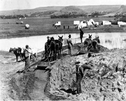 Black and white photo of men and horses working near a river, with covered wagons and tents in the background.