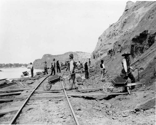 Black and white photo of workers with wheelbarrows near railroad tracks, surrounded by rocky terrain and a river.
