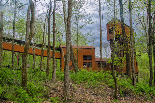 Abandoned industrial structure surrounded by trees, with a rusty tower and conveyor belt amidst lush greenery.