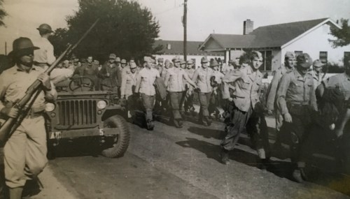 Black and white photo of soldiers marching down a street, with a jeep and houses in the background.
