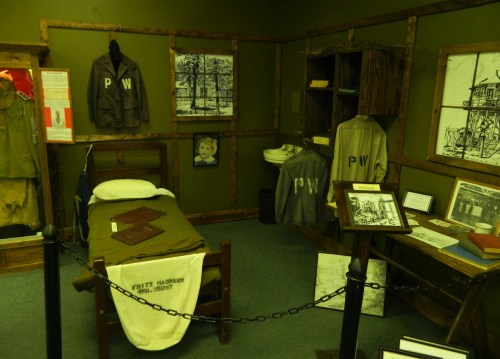 A historical exhibit featuring a prisoner's room with clothing, a bed, and various artifacts displayed on walls and stands.