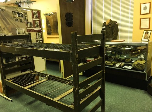 A military museum display featuring a bunk bed and a showcase of historical uniforms and artifacts.
