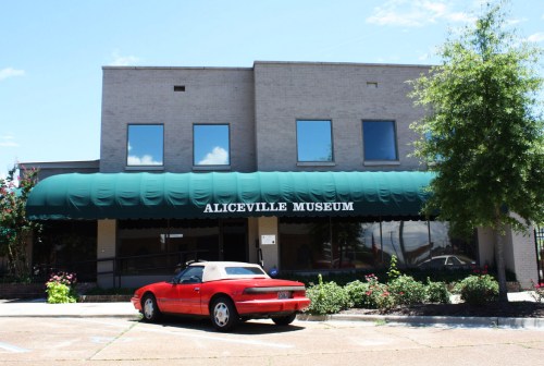 Exterior of the Aliceville Museum, featuring a green awning and a red convertible parked in front.