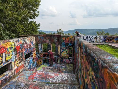 Two people explore a colorful, graffiti-covered ruin with a scenic view of hills in the background.