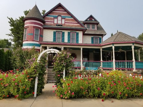 Colorful Victorian house with a turret, surrounded by vibrant flower beds and a decorative archway.