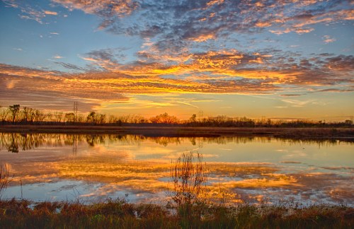 A serene sunset over a calm lake, with vibrant clouds reflecting on the water and silhouettes of trees in the background.