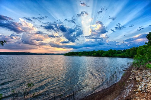 A serene lake at sunset, with vibrant clouds and rays of light illuminating the sky above lush greenery along the shore.