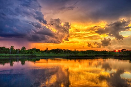 A vibrant sunset over a calm lake, with dramatic clouds reflecting in the water and lush greenery in the background.